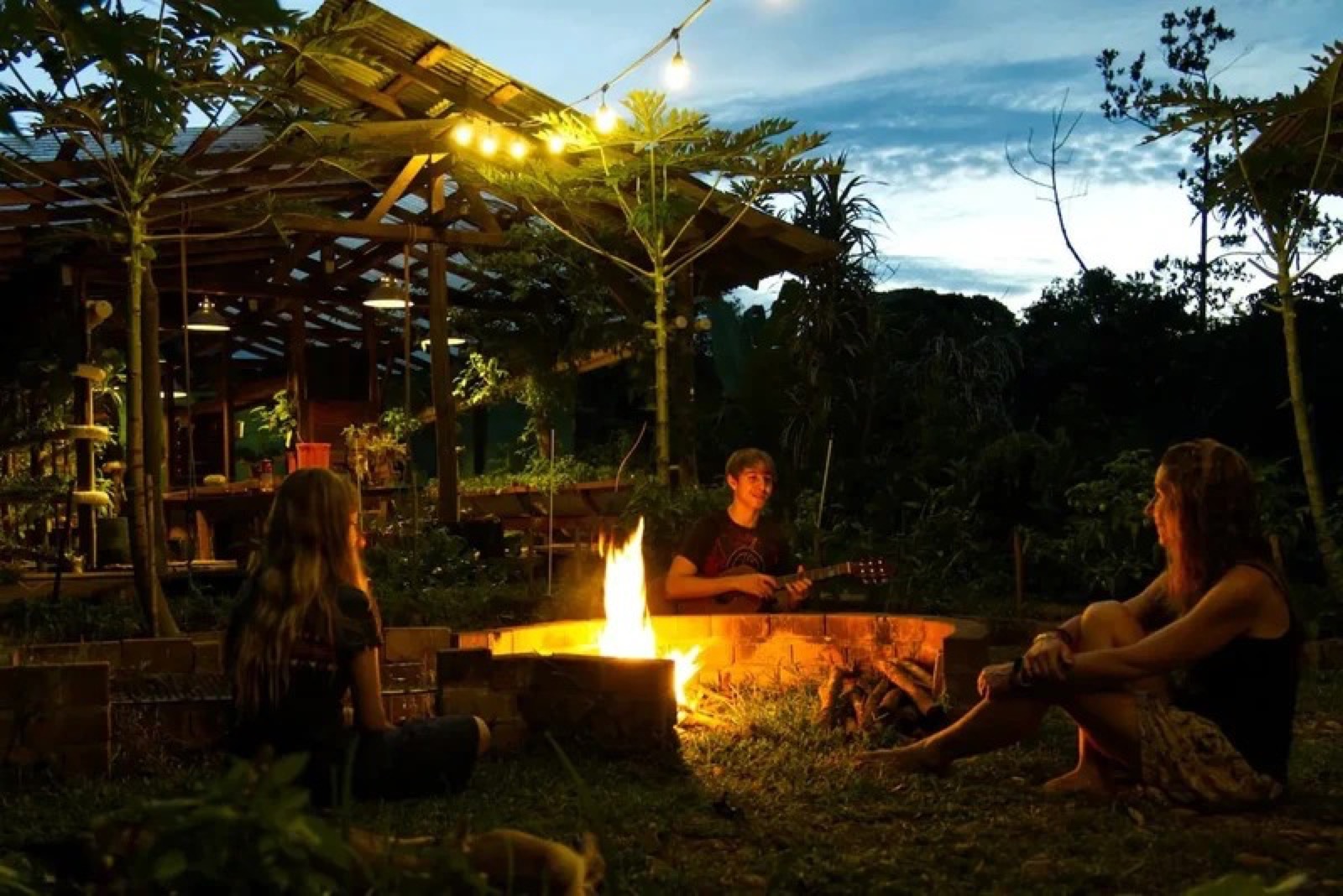 Friends around the fire pit at dusk beneath the string-lit pavilion